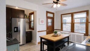 Una cocina con refrigerador, estufa, mesa de madera, ventilador de techo y ventanas.