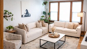 Una sala de estar con dos sofás beige, un sillón estampado, una mesa de centro y un ventanal con persianas.
