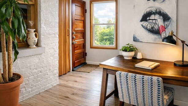 Una habitación con puerta de madera, ventana con vistas, escritorio con lámpara, una silla y una planta en maceta.