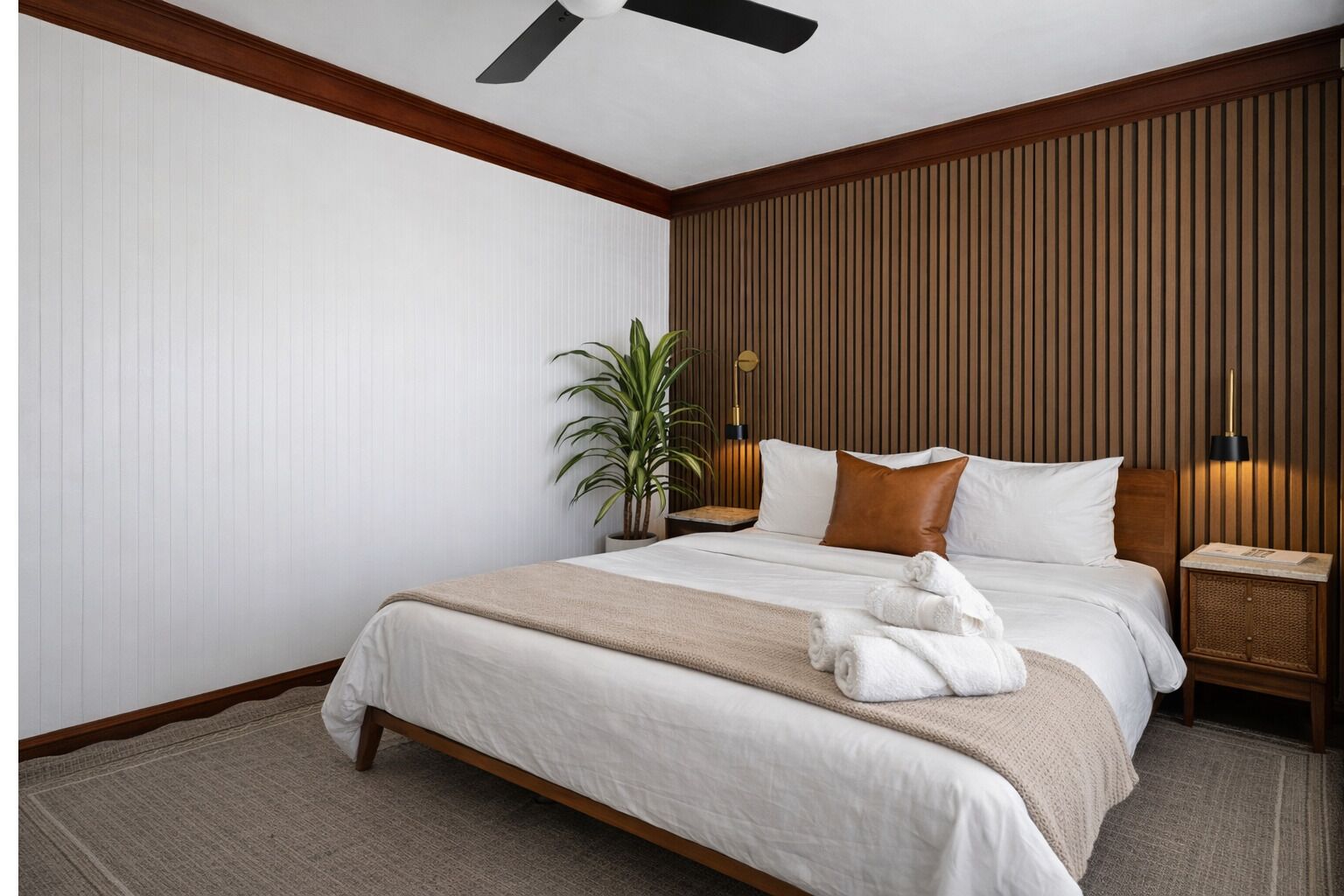 Una habitación de hotel con cama, ventilador de techo, mesita de noche, lámpara y planta en maceta.