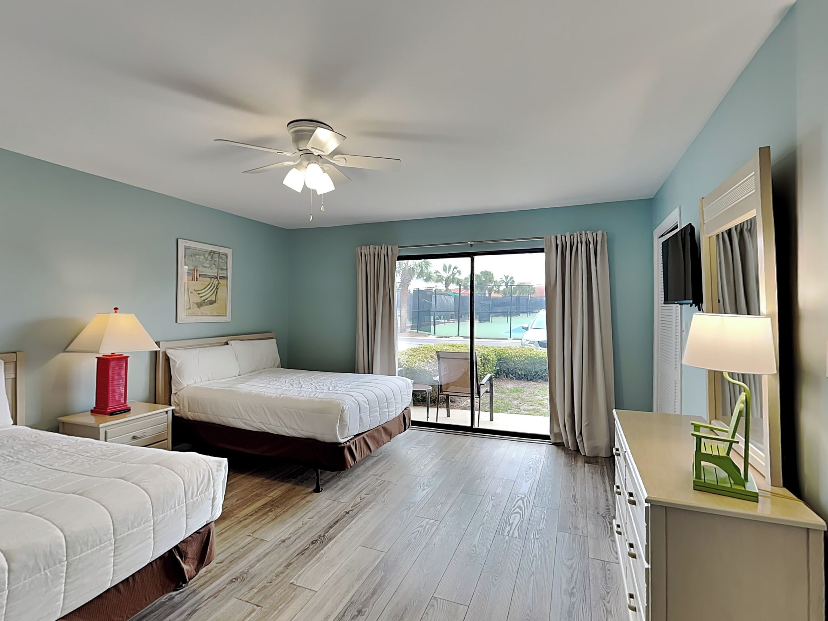 Habitación de hotel con cama, dos mesitas de noche con lámparas, ventilador de techo, ventana grande con cortinas y vista a una cancha de tenis.