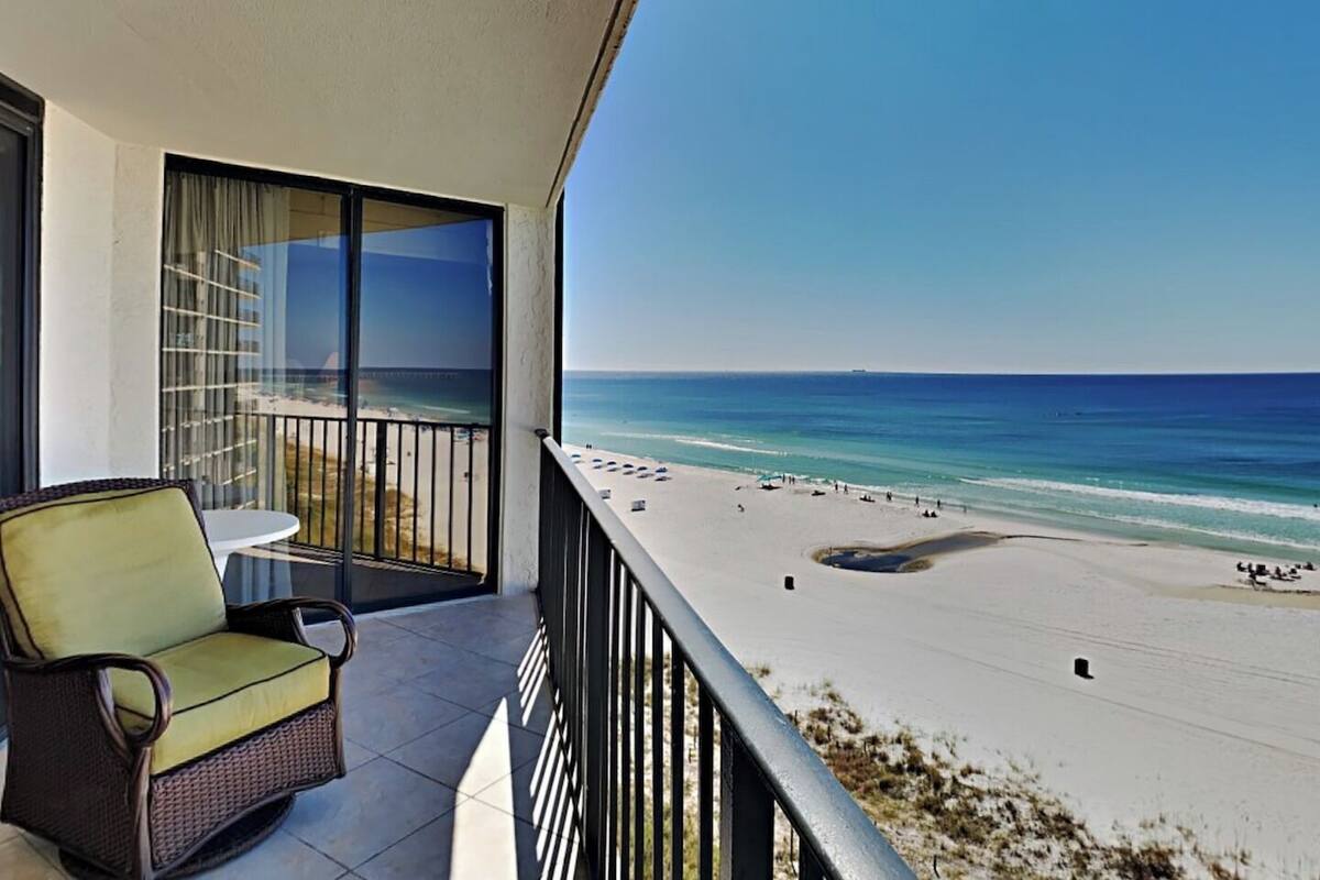 Een balkon met een rieten stoel, een glazen deur en uitzicht op een strand met mensen en een heldere lucht.