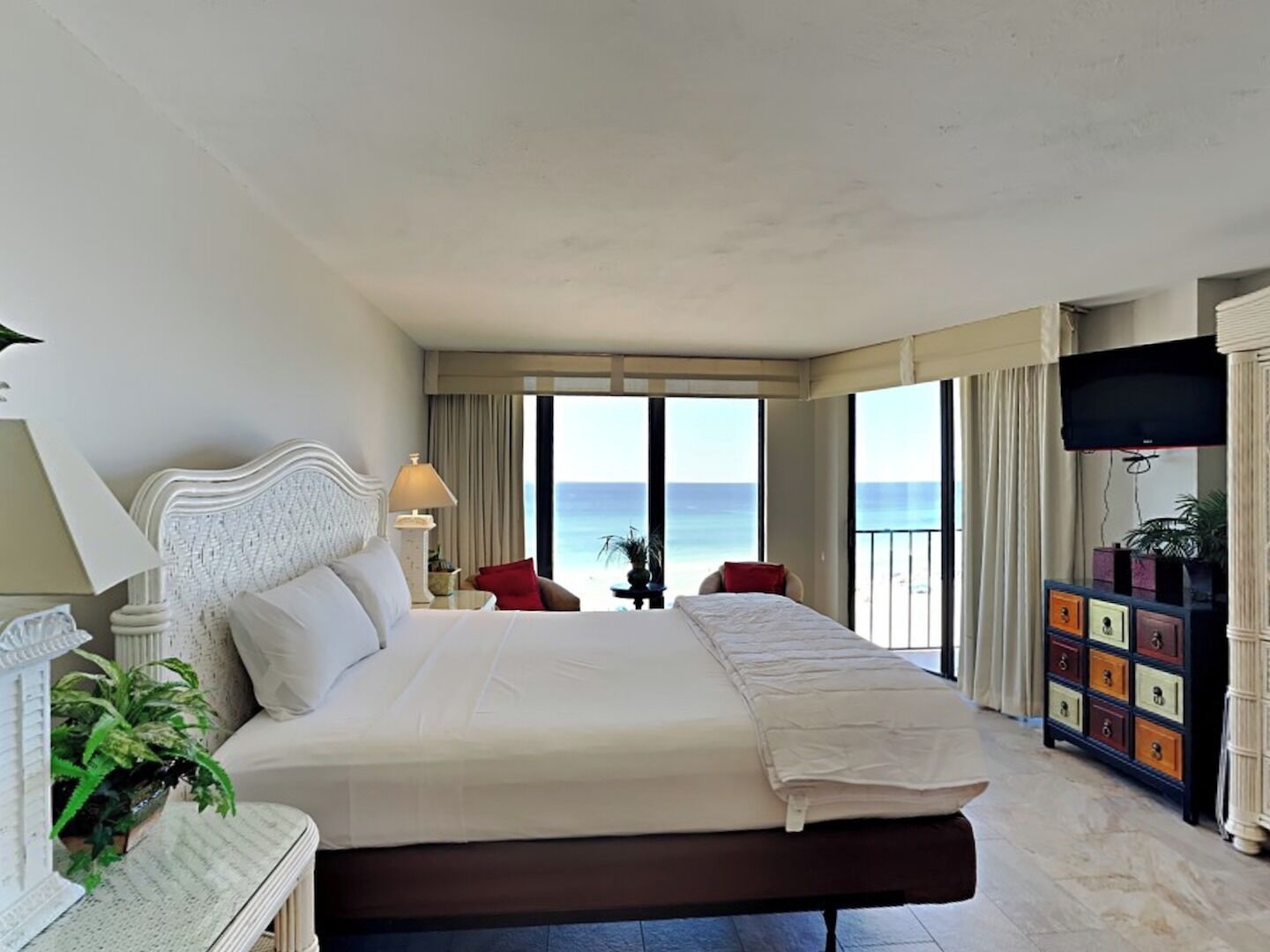 A hotel room with a large bed, a TV, a bedside table with a lamp, a plant, and a view of the ocean.