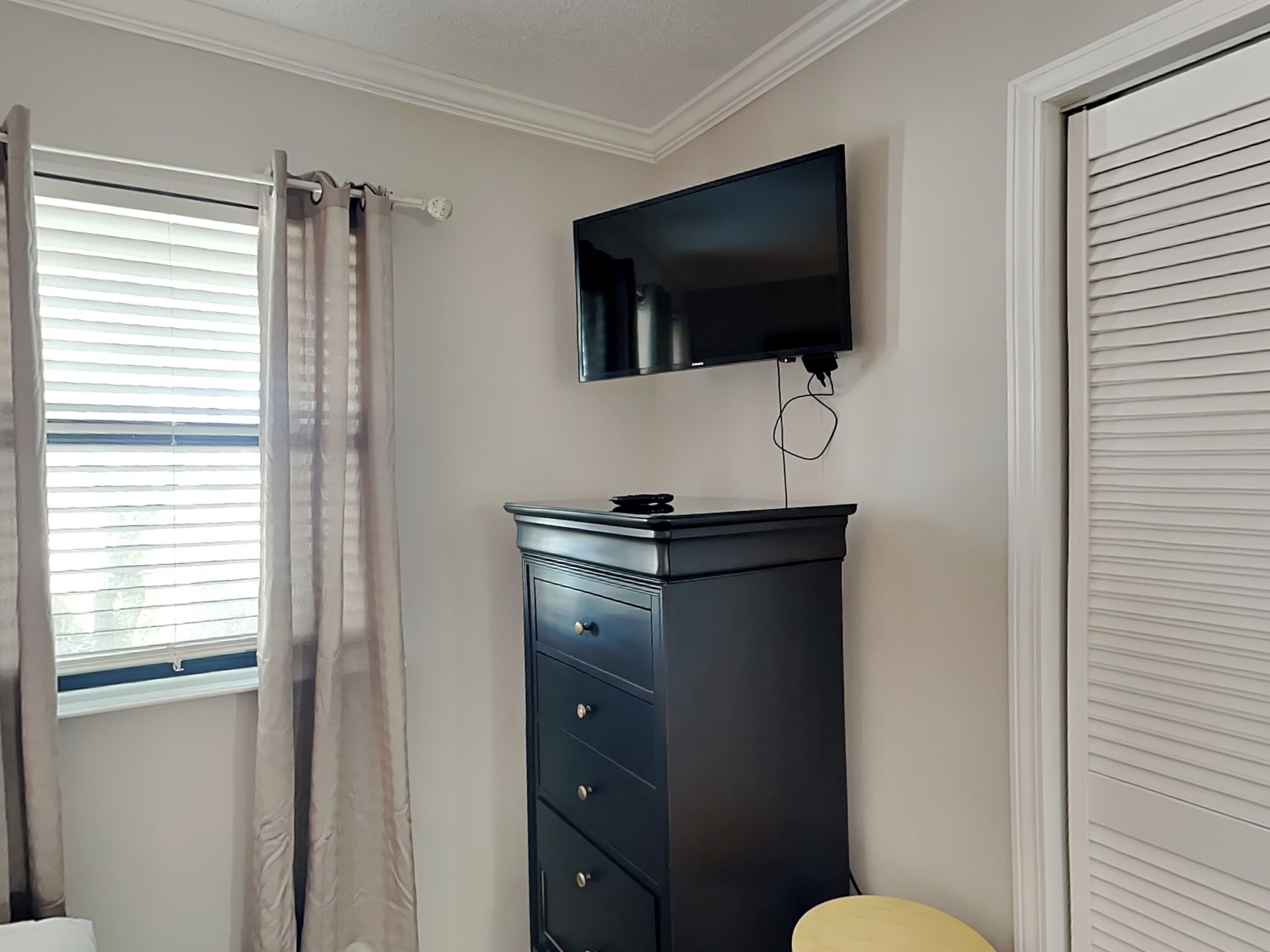 A room with a window, a black dresser, a television mounted on the wall, and a closed door.