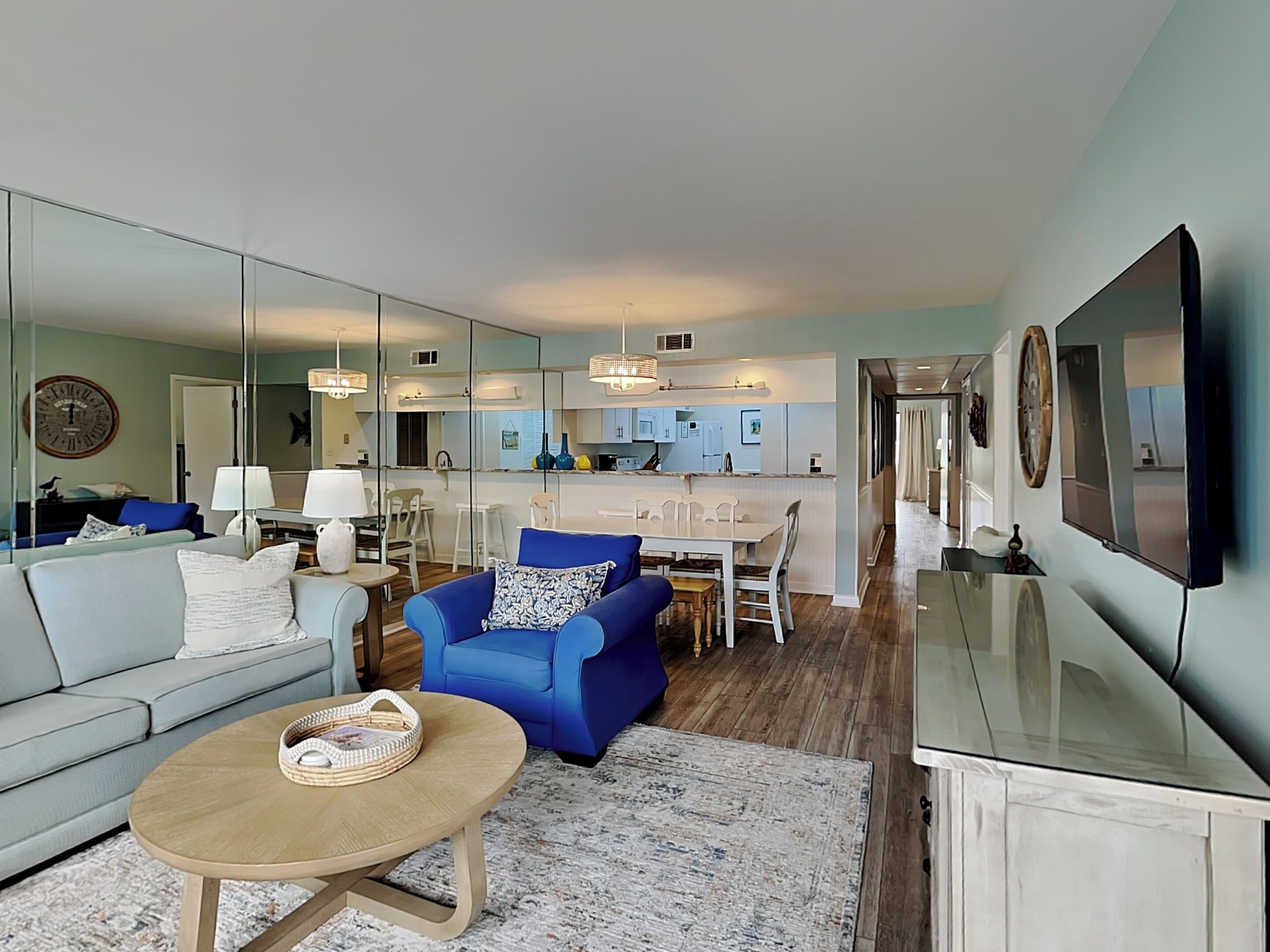 Un salon moderne avec un canapé, un fauteuil bleu et une table basse. On aperçoit à l’arrière-plan un coin cuisine avec une table à manger et des chaises.