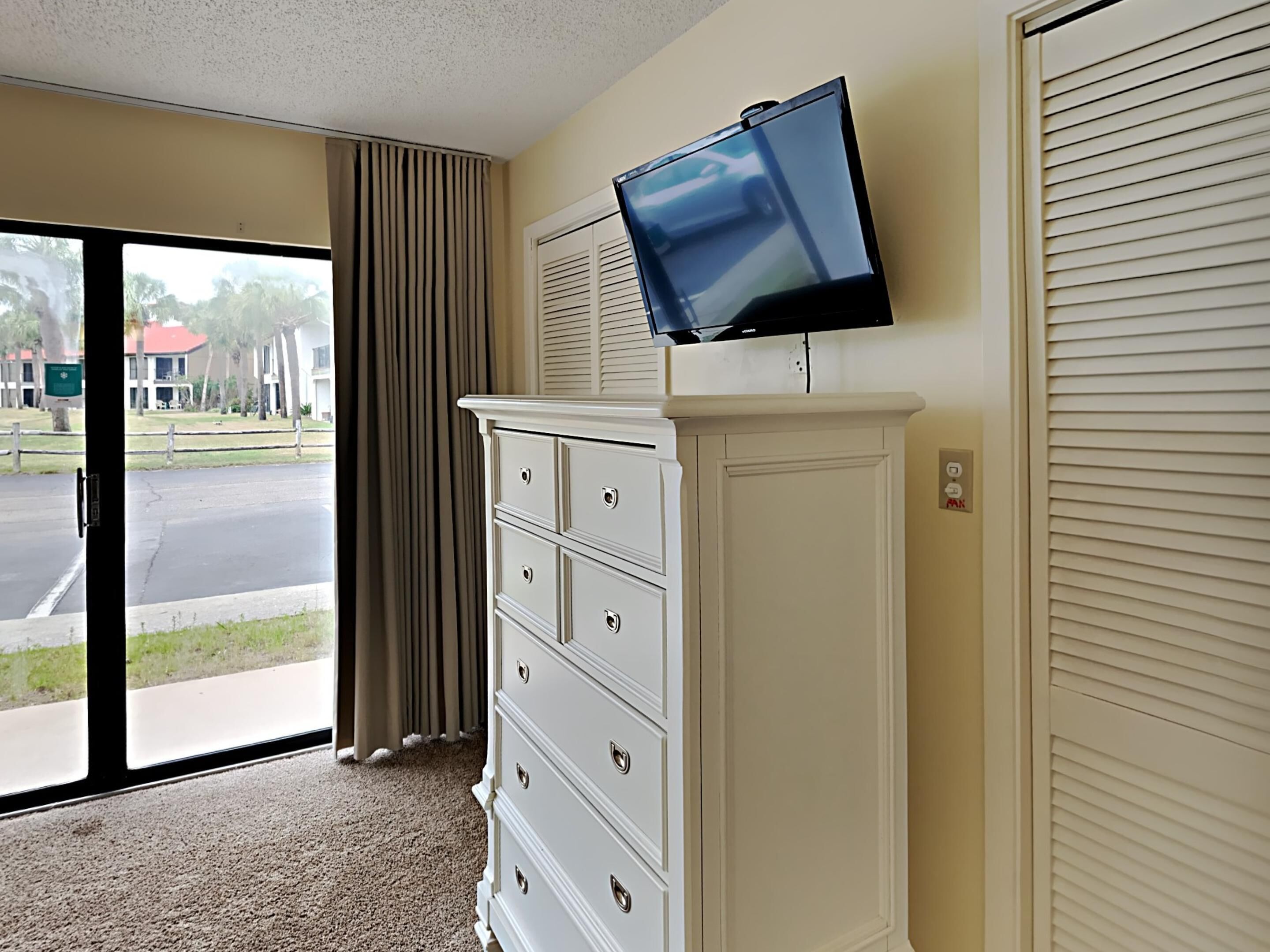 Ein Hotelzimmer mit großem Fenster, einem weißen Schminktisch, einem flachbildfernseher, der darüber angebracht ist, und einer geschlossenen Schiebetür.