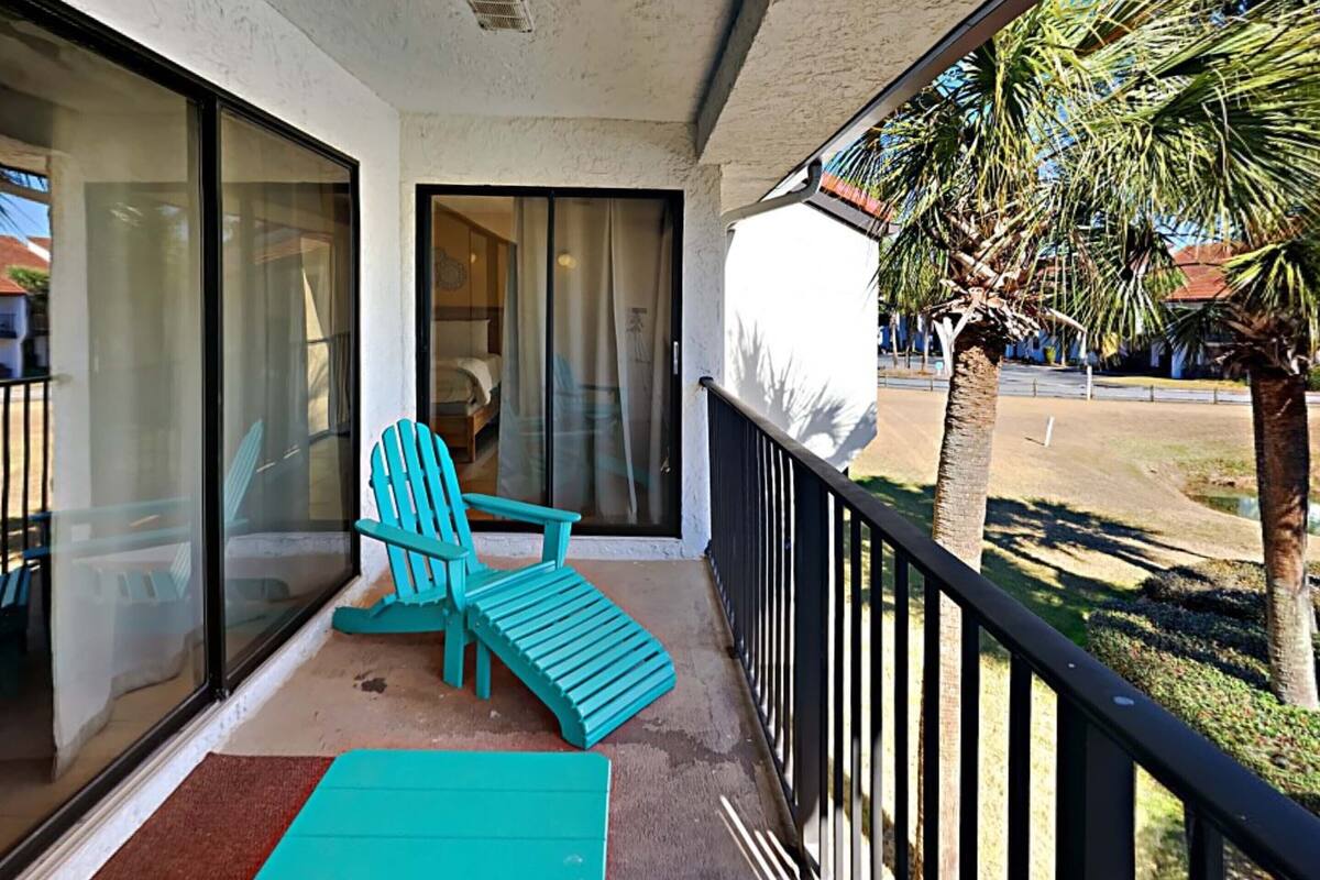 Een balkon met een blauwgroene stoel en een klein tafeltje, een glazen schuifdeur en een palmboom op de achtergrond.
