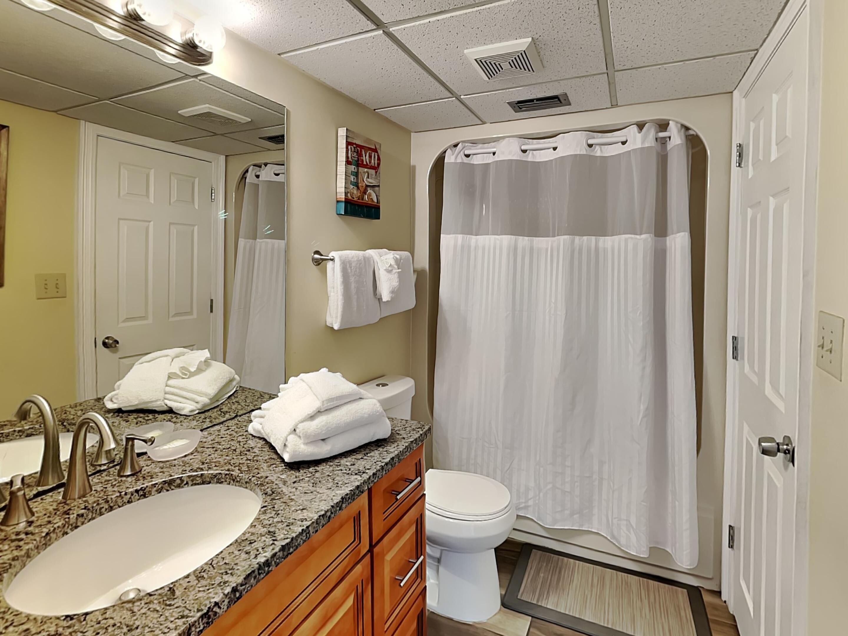 Une salle de bain avec un lavabo sur un plan de travail en granit, des toilettes blanches, une douche avec un rideau blanc et un porte-serviettes avec des serviettes pliées.