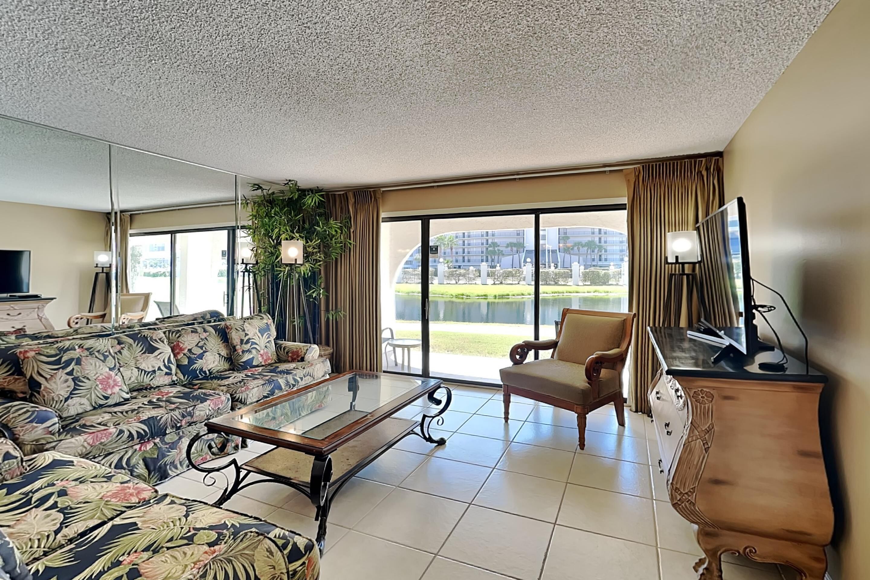 A living room with a sofa, armchair, and coffee table, large windows with a view of a building and greenery, and a flat-screen TV on a wooden cabinet.