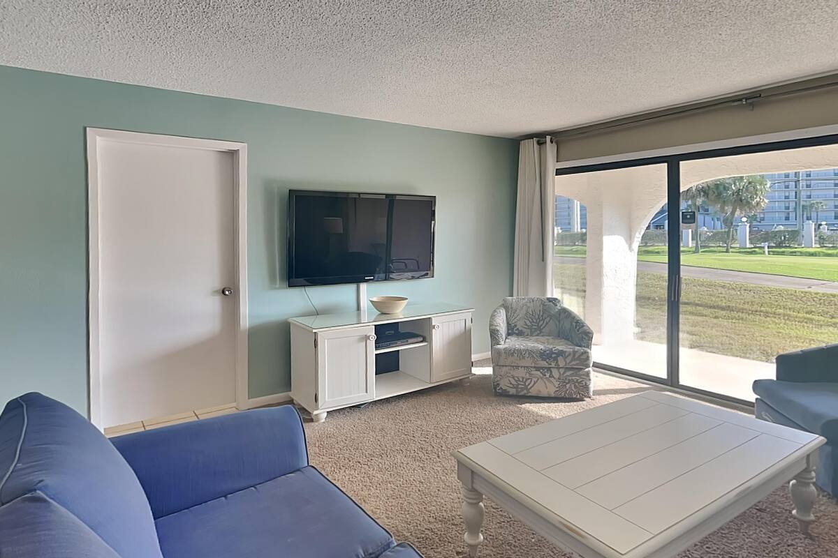 Een woonkamer met een grote flatscreentelevisie, een witte tv-kast, een blauwe bank, een fauteuil met een patroon en uitzicht op een groen gazon door een glazen deur.