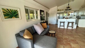 A living room with a grey sofa, a small round table, and a kitchen area with a white island and stools. - Ylang Ylang Luxury 2 Suite Villa - Cruz Bay, St. John (Cruz Bay)