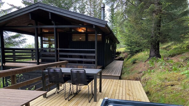 A wooden deck with a hot tub, a table with chairs, and a covered seating area with a view of the surrounding forest. - The Secret Cabin with hot tube in nature reserve close to Reykjavik  (Hafnarfjordur)