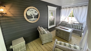 Eine überdachte Veranda mit dunkel verkleideter Fassade, Korbmöbeln, einer an der Wand befestigten Baumdekoration und einem Fenster mit weißen Vorhängen.
