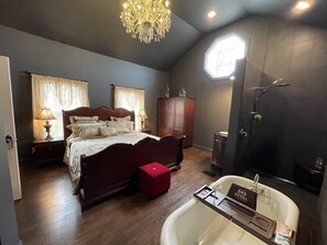 A spacious bedroom with a large bed, a wooden wardrobe, a red ottoman, and a bathroom with a bathtub and a vanity.