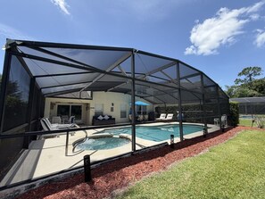 A pool area with a covered seating area, a hot tub, and a house in the background.