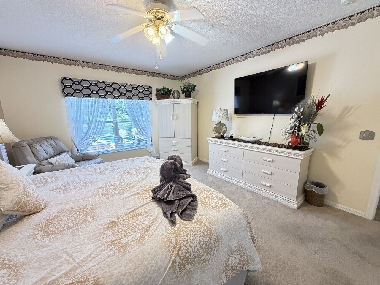 Un dormitorio con cama, ventilador de techo, televisor, cómoda y ventana con cortinas.