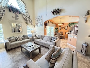 A spacious living room with a sofa set, coffee table, and decorative pillows. A kitchen area is visible in the background. - Luxury Lakeside Pool & Spa Home close to Disney (Kissimmee)