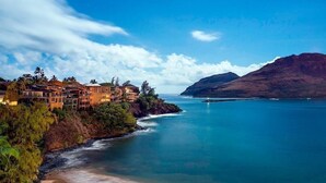 A coastal hotel with multiple buildings, a beach, and a mountain in the background. - Modern Oceanfront Resort Living. Luxury Oceanfront Bedroom Suites. Sleeps 10! (Lihue)