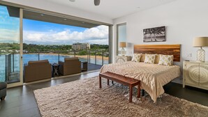 A bedroom with a large bed, a wooden headboard, a view of the sea, and a balcony with outdoor seating. - Modern Oceanfront Resort Living. Luxury Oceanfront Bedroom Suites. Sleeps 10! (Lihue)