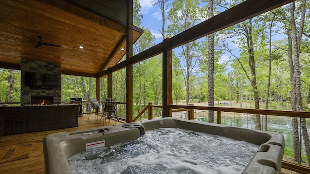 A hot tub in a wooden deck area with a stone fireplace and outdoor furniture. - Couple's Escape + King Bed + Fountain + Hot Tub (Broken Bow)