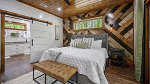 A bedroom with a large bed, a bench, a dresser, and a window with a view of trees. - Couple's Escape + King Bed + Fountain + Hot Tub (Broken Bow)