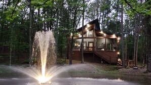 A cabin with a large window, a wooden deck, and a waterfall feature in front. - Couple's Escape + King Bed + Fountain + Hot Tub (Broken Bow)