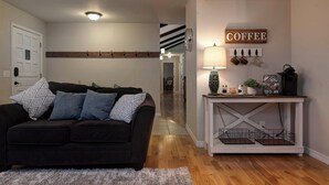 A living room with a dark brown sofa, a white door, a wooden bench, a coffee-themed wall decoration, and a lamp on a white console table. - Family Fun at Primrose Retreat - Cozy, Clean & Chemical-Free (Republic)