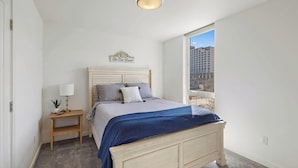 A bedroom with a bed, bedside table, lamp, and a view of buildings through a window.