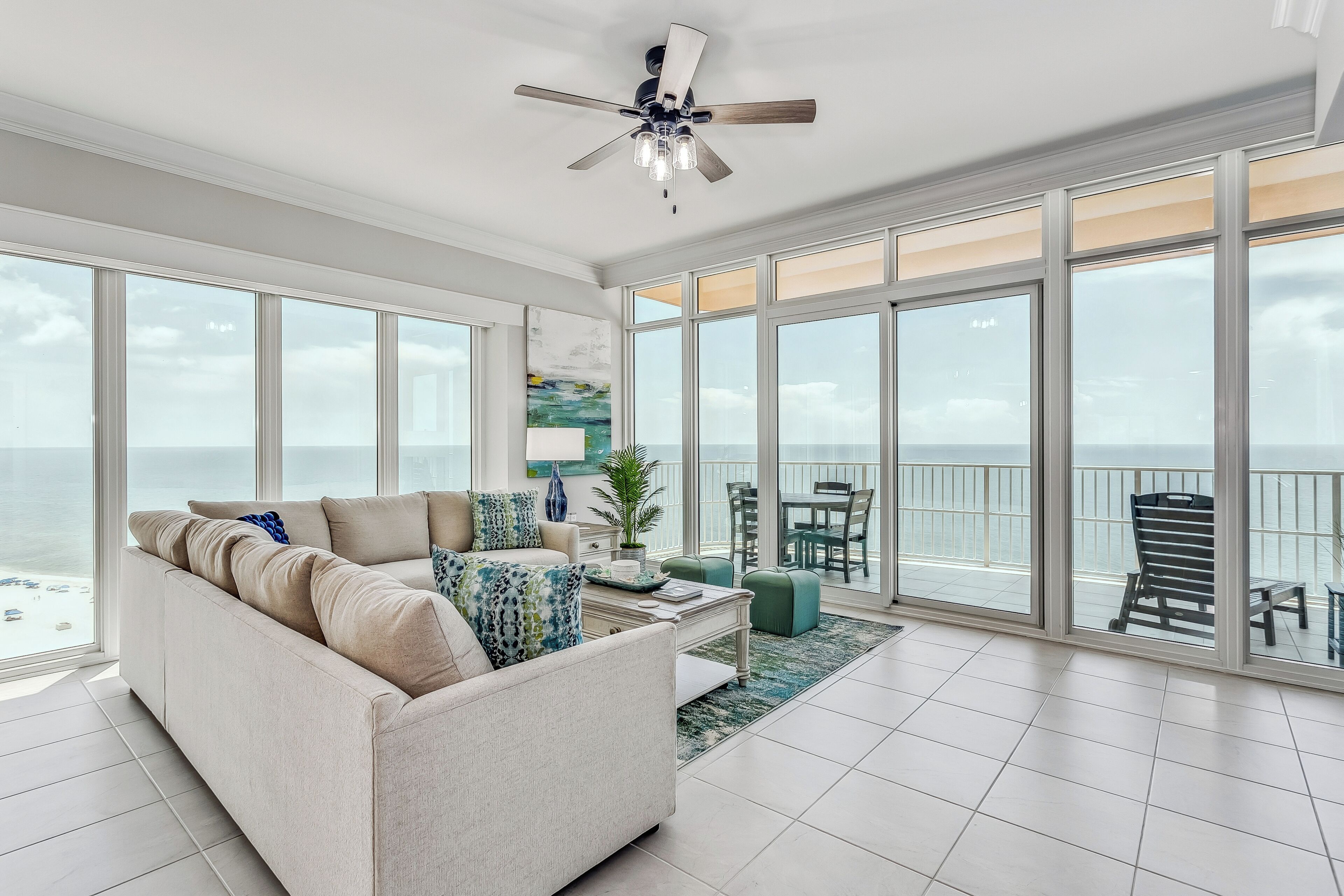 A spacious living room with a large window overlooking the ocean, a ceiling fan, and a dining area with outdoor seating.