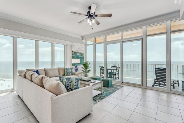 A spacious living room with a large window overlooking the ocean, a ceiling fan, and a dining area with outdoor seating.