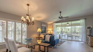 A spacious living area with a glass dining table, white chairs, a blue sofa, and a ceiling fan.