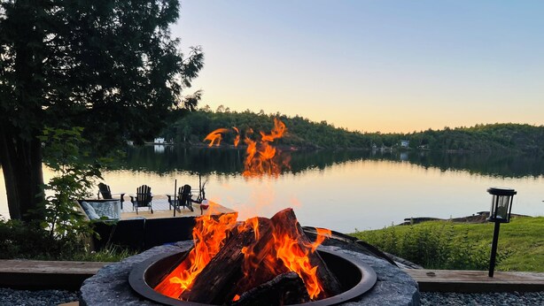 A circular stone fire pit with flames, surrounded by pebbles, overlooking a calm lake with docked chairs and a lamp post. - 5-Bedroom Executive Lakefront Retreat Sleeping 12- Relax, Recharge and Reconnect (Greater Sudbury)