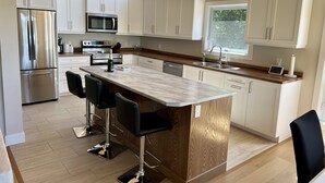 A modern kitchen with white cabinets, stainless steel appliances, a wooden island with bar stools, and a window providing natural light. - 5-Bedroom Executive Lakefront Retreat Sleeping 12- Relax, Recharge and Reconnect (Greater Sudbury)