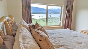Una cama con muchas almohadas y vistas a un lago y montañas a través de una ventana.