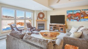 Amplia sala de estar con chimenea, ventanas grandes con vistas a la montaña, ventilador de techo y televisor montado.