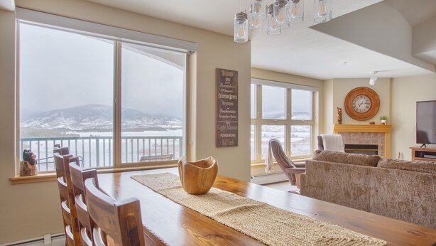 Un comedor con una mesa y sillas de madera, un ventanal con vistas a montañas y una chimenea con un reloj en la pared.