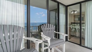 Ein Balkon mit zwei Adirondack-Stühlen, einer Glastür, die zu einem Zimmer führt, und Blick auf den Ozean.