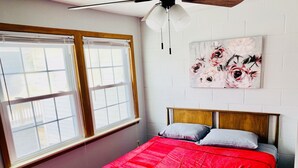 A bedroom with a bed, a ceiling fan, a window with blinds, and a floral painting on the wall.