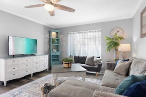 Un salon comprenant une télévision, une commode blanche, une étagère turquoise, un canapé gris, une table basse et un lampadaire.