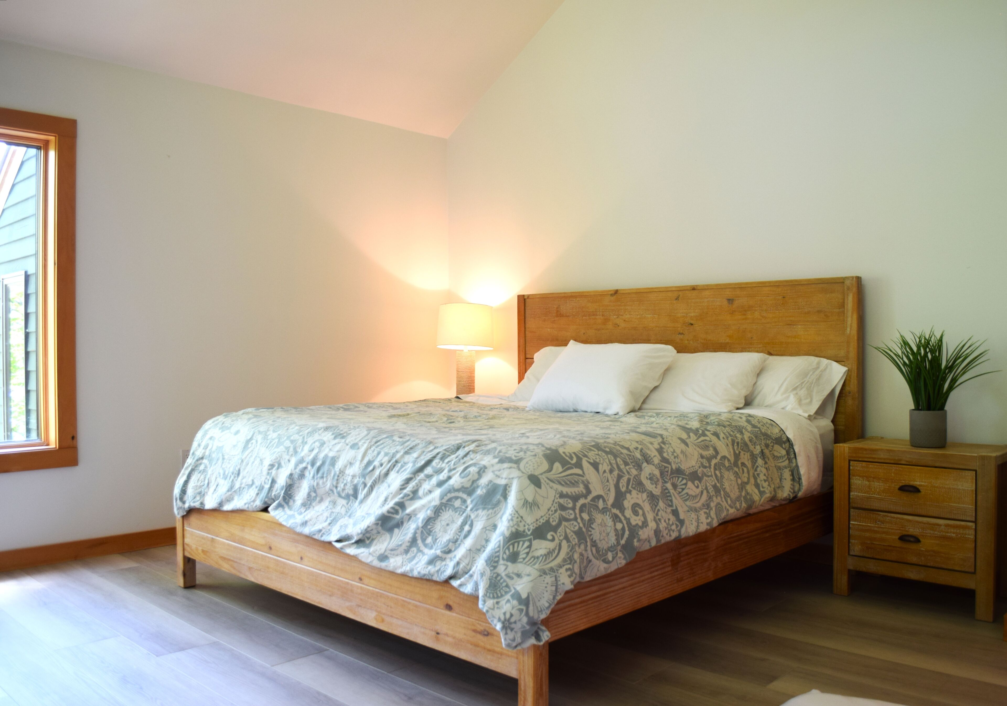 Une chambre à coucher avec un lit en bois, une lampe de chevet, une table de nuit ornée d’une plante et une fenêtre avec des stores.