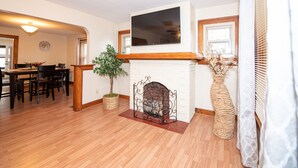 Ein Wohnzimmer mit Kamin, Holzboden, einer Essecke mit Tisch und Stühlen sowie einem großen Flachbildfernseher, der über dem Kamin angebracht ist.