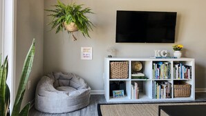 Ein Wohnzimmer mit grauem Sitzsack, einem weißen Bücherregal, einer hängenden Pflanze und einem Flachbildfernseher an der Wand.