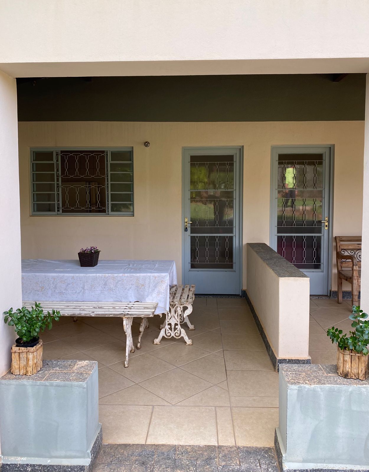 A covered outdoor seating area with a table and benches, two doors with metal bars, and potted plants.
