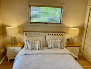 A bedroom with a bed, two bedside tables with lamps, a window with blinds, and a small plant on one table.