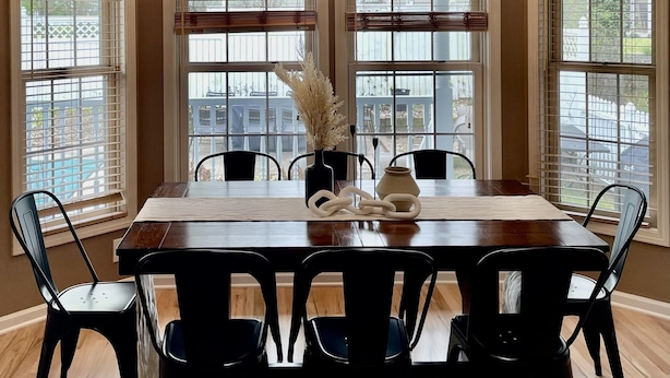 Une salle à manger avec une table en bois, des chaises noires, un vase contenant des fleurs séchées et une horloge murale.