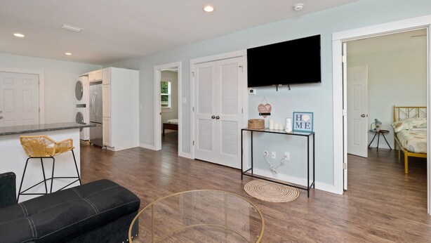 A modern kitchen with white cabinets, a built-in oven, and a refrigerator. There is a dining area with a black couch and a wicker chair. A flat-screen TV is mounted on the wall, and there is a small table with a basket and candles. - Stylish 2 bedroom home close 2 Dtown, NAS, and BCH (Pensacola)
