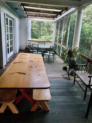 A covered outdoor dining area with a long wooden table and chairs, surrounded by windows and plants.