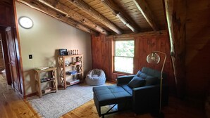 Quarto aconchegante com teto de madeira, sofá, puff, estante de livros e janela com vista para o verde.