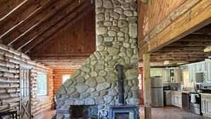 Interior rústico de cabana com lareira de pedra, vigas de madeira, ventilador de teto e área de jantar com mesa e cadeiras.