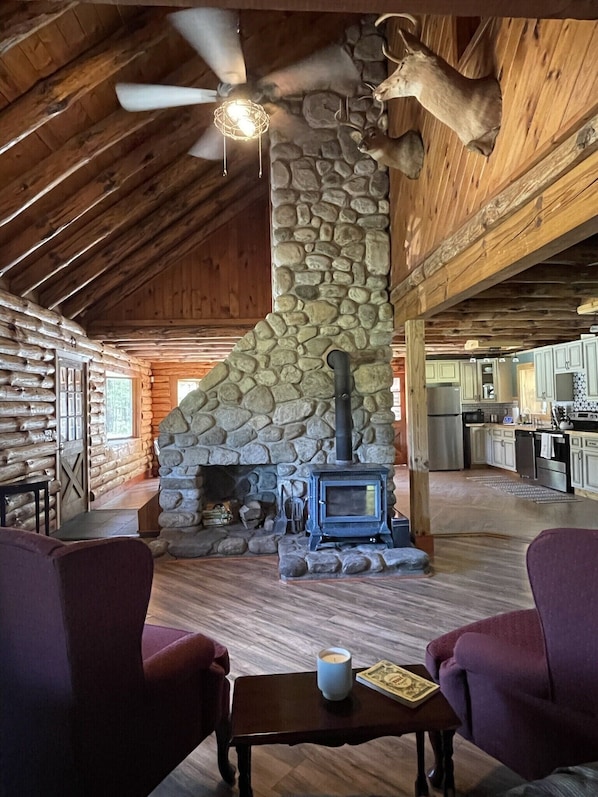 Interior rústico de cabana com lareira de pedra, vigas de madeira, ventilador de teto e área de jantar com mesa e cadeiras.